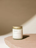 A glass jar candle labeled "MINOT" and "143" with notes of mandarin, sandalwood, and jasmine sits on a stone surface. The lid is golden, and soft lighting creates gentle shadows.