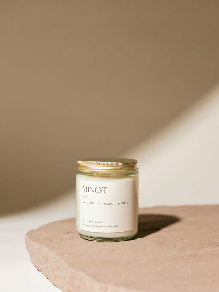 A glass jar candle labeled "MINOT" and "143" with notes of mandarin, sandalwood, and jasmine sits on a stone surface. The lid is golden, and soft lighting creates gentle shadows.