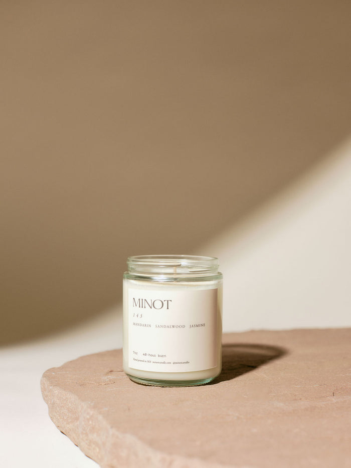 A glass jar candle labeled "MINOT" and "143" (for "I Love You") with scents of mandarin, sandalwood, and jasmine is placed on a textured stone surface against a beige background.
