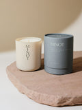 A non-toxic soy candle in a glass vessel labeled "MINOT" sits on a textured stone surface next to a gray cylindrical box labeled "Aeris: Sandalwood, Lavender, Fresh Citrus"