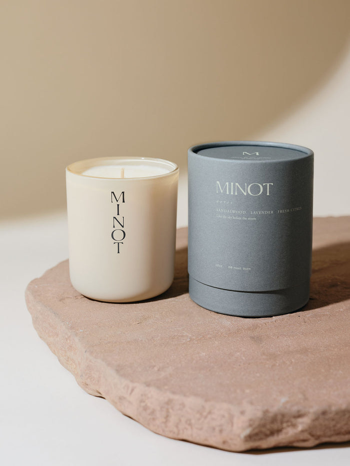 A non-toxic soy candle in a glass vessel labeled "MINOT" sits on a textured stone surface next to a gray cylindrical box labeled "Aeris: Sandalwood, Lavender, Fresh Citrus"