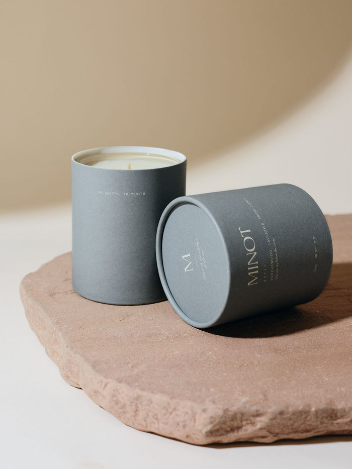 Blue-gray cylindrical packaging holds a MINOT Aeris scented candle with its lid tilted to the side on a textured stone slab. The background is a soft beige color.