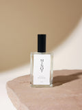 An elegant glass bottle of MINOT Aeris Room & Linen Spray sits on a textured stone surface. The label is white with black text, and the cap is black. The background is softly lit in neutral tones.