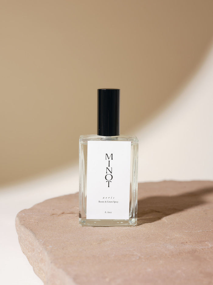 An elegant glass bottle of MINOT Aeris Room & Linen Spray sits on a textured stone surface. The label is white with black text, and the cap is black. The background is softly lit in neutral tones.