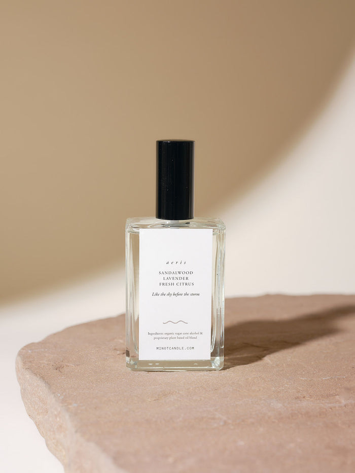 A glass bottle of MINOT Aeris Room & Linen Spray sits on a textured stone surface. The label reads "Aeris: Sandalwood, Lavender, Fresh Citrus" and "Like the sky before the storm."