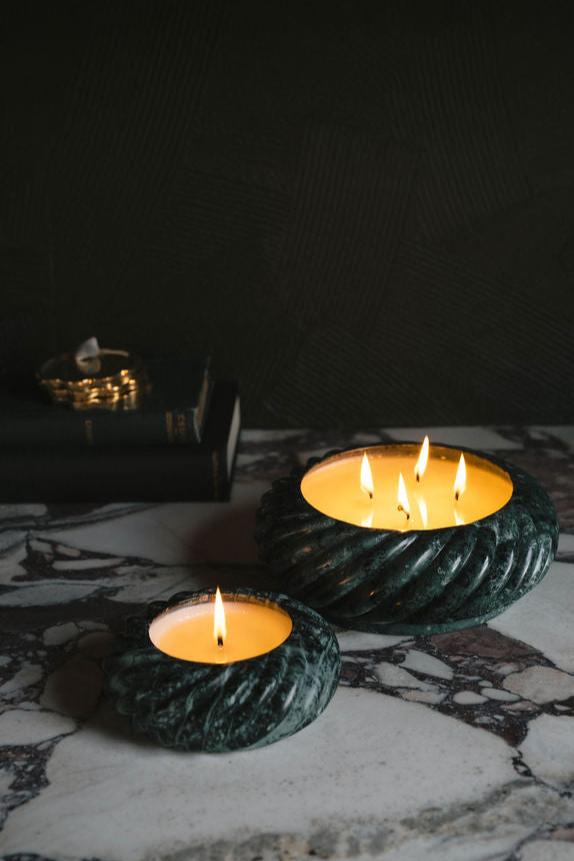 Two dark green stone vessels holding lit candles sit on a marble surface.