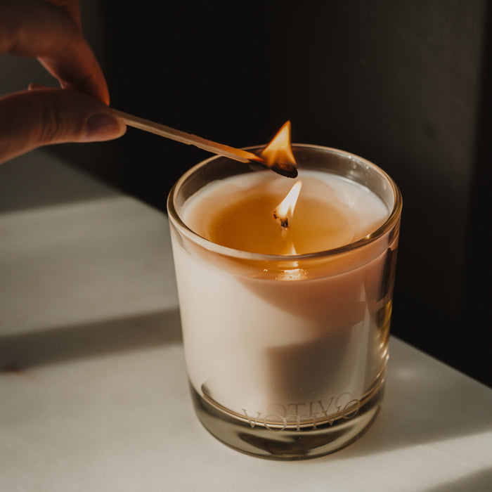 A hand lights the Votivo Bamboo Leaf 10 oz. Signature Candle, made from Soy Blend Wax with a Bamboo Leaf Fragrance, in a glass holder on a white surface as the flame ignites the wick.