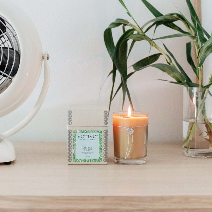A Votivo Bamboo Leaf 6.8 oz. Aromatic Candle, a small box labeled “Votivo Bamboo Leaf,” a green plant in a glass, and a white fan are arranged on a light wooden surface against a white wall.