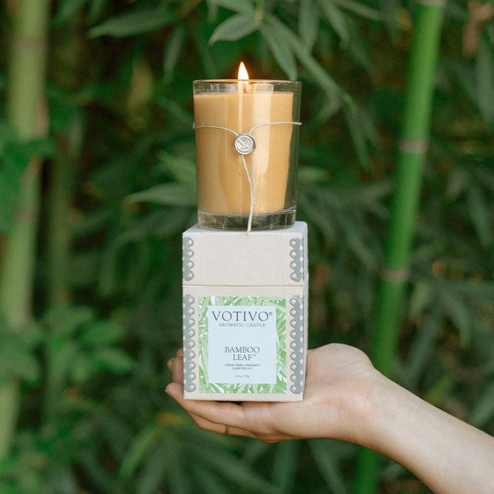 A hand holds a box labeled Votivo Bamboo Leaf 6.8 oz. Aromatic Candle with a lit candle on top, set against lush green bamboo plants.