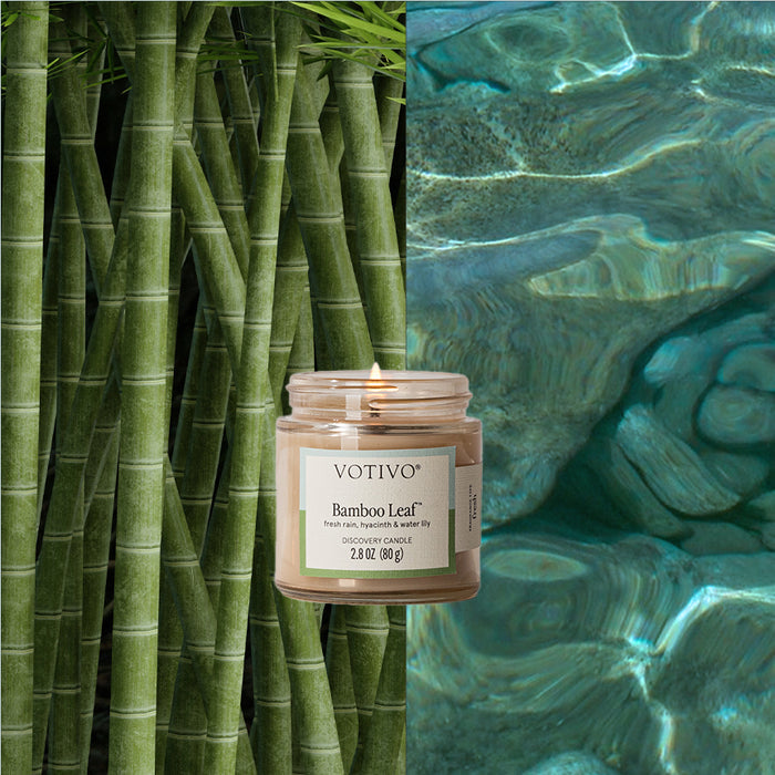 A Votivo Bamboo Leaf Discovery Candle burns in a glass jar, set between close-up green bamboo stalks on the left and clear, rippling blue water on the right.