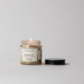 A lit Votivo Bamboo Leaf Discovery Candle rests in a glass jar on a white surface, its black lid beside it. The label is visible against a light background, highlighting the soy blend wax and fresh fragrance.