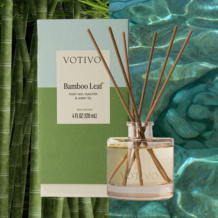 A Votivo Bamboo Leaf Reed Diffuser with several fiber reeds in a clear glass bottle rests before its green and blue box, set against a serene scene of bamboo stalks and water.