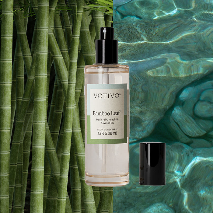 The Votivo Bamboo Leaf Room & Linen Spray, shown with its black cap off, is set before bamboo stalks and a clear blue pool, reflecting the products fresh scent.