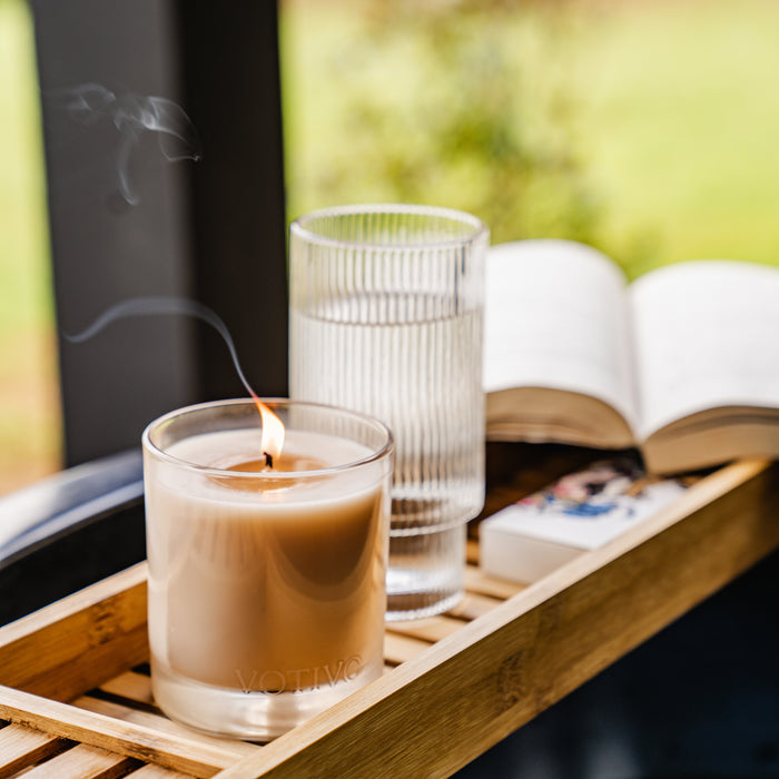 A Votivo Champaca 10 oz. Signature Candle, a glass of water, an open book, and a matchbox sit on a wooden tray outdoors amid blurred greenery—a tranquil scene elevated by the candle’s unique fragrance.