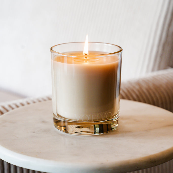 The Votivo Champaca 10 oz. Signature Candle glows in a clear glass holder on a round white marble table, with a beige upholstered chair in the background.