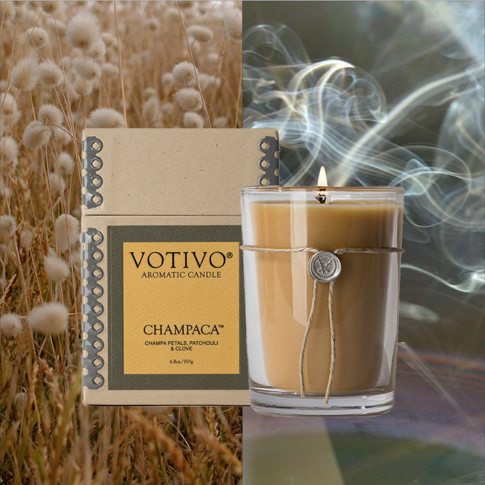 The Votivo Champaca 6.8 oz. Aromatic Candle and its box are displayed among soft grasses. The lit soy blend wax candle, adorned with a decorative string and seal, sits beside the box with swirling smoke in the background.