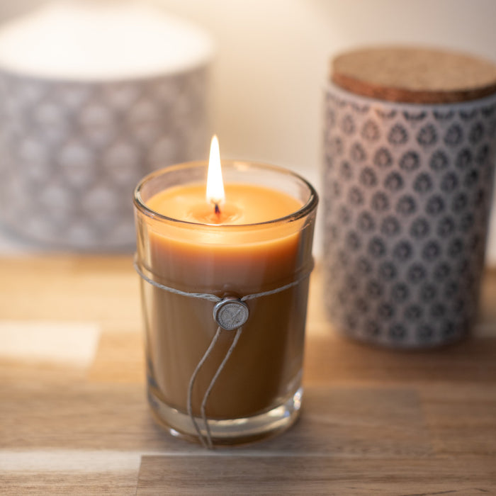 The Votivo Champaca 6.8 oz. Aromatic Candle, made with a soy blend and set in a twine-wrapped glass holder on wood, creates a warm patchouli-inspired ambiance as two patterned containers blur softly in the background.