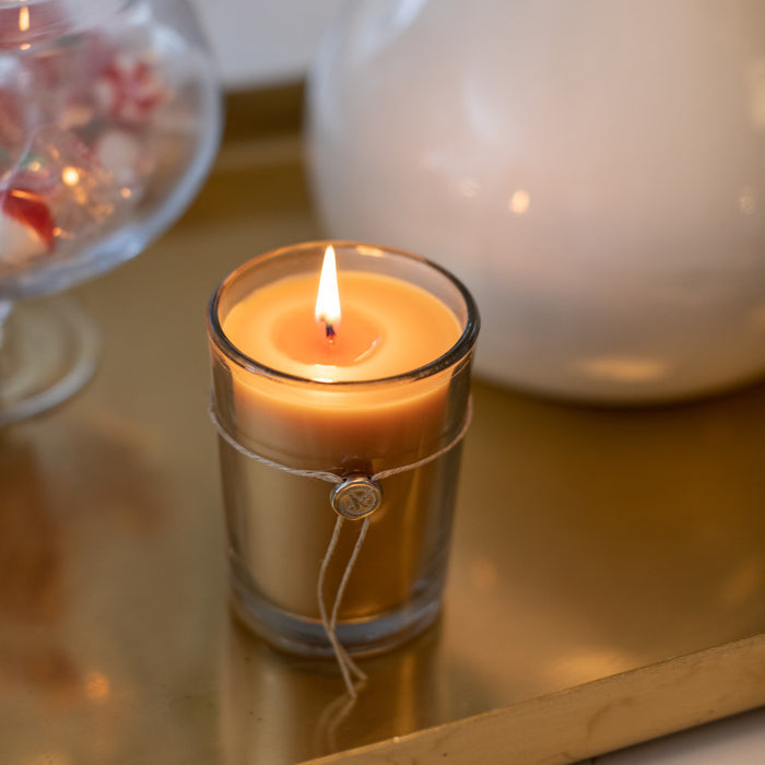 The Votivo Champaca 6.8 oz. Aromatic Candle, made from soy blend wax and set in a glass holder with a wax seal and string, rests on a gold tray. A white vase and a glass bowl of red and white candies are blurred behind it.