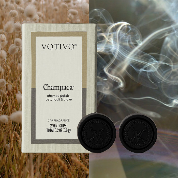 A Votivo Champaca Car Fragrance (2 Pack) is centered with two black vent clips; beige pampas grass on the left and swirling smoke on the right evoke patchouli’s woody scent against a dark background.