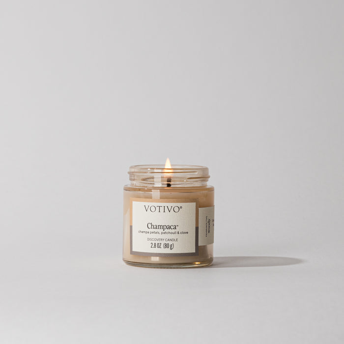 A Votivo Champaca Discovery Candle with beige soy-blend wax burns with a single flame against a plain light gray background, featuring a minimalist design and filling the air with its subtle woody scent.