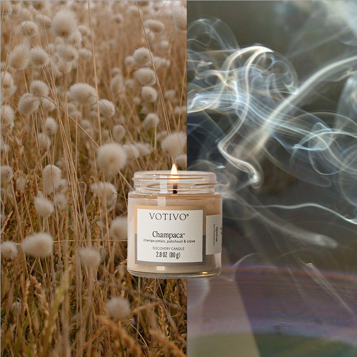 A glowing Votivo Champaca Discovery Candle is centered, paired with soft field plants on the left and curling smoke on the right, creating a scene that highlights its inviting woody fragrance and natural appeal.