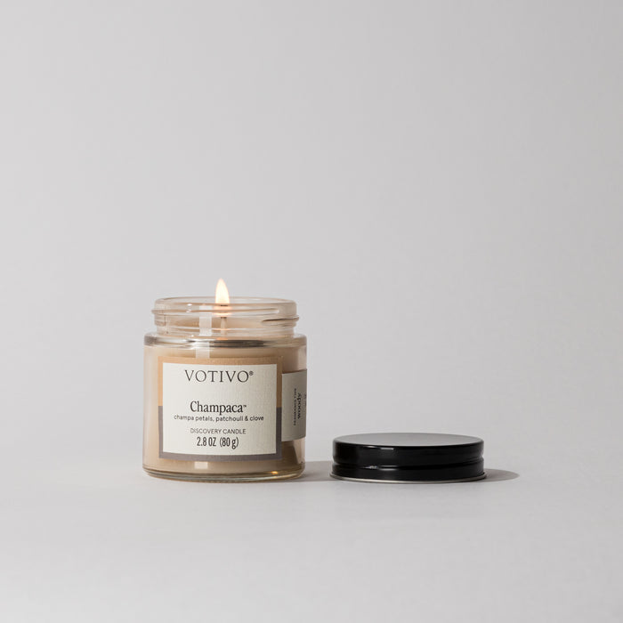 A Votivo Champaca Discovery Candle with a soy blend sits in its glass jar beside the black lid on a plain, light background. The front label is visible, suggesting the candle’s woody fragrance.