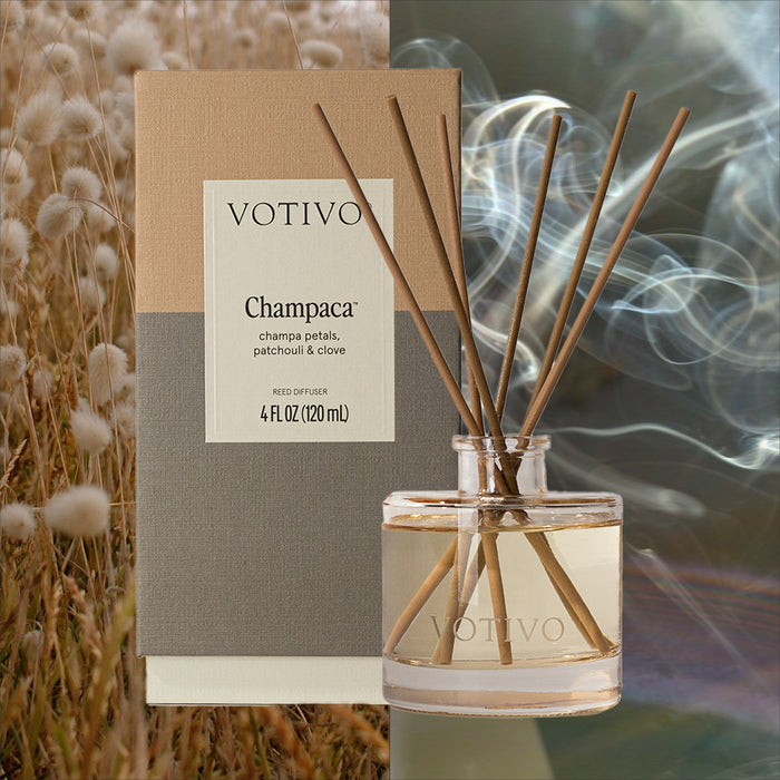 A Votivo Champaca Reed Diffuser with fiber reeds and a woody scent is displayed in a glass jar before its box, set against soft beige plants to the left and swirling smoke to the right.