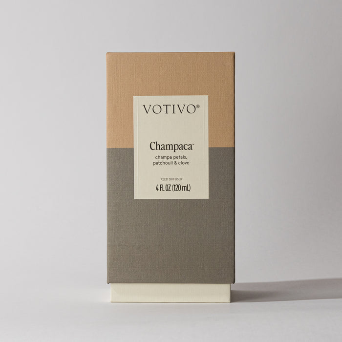 A Votivo Champaca Reed Diffuser box, beige and gray, displays a woody scent with champa petals, patchouli, and clove notes; fiber reeds included.