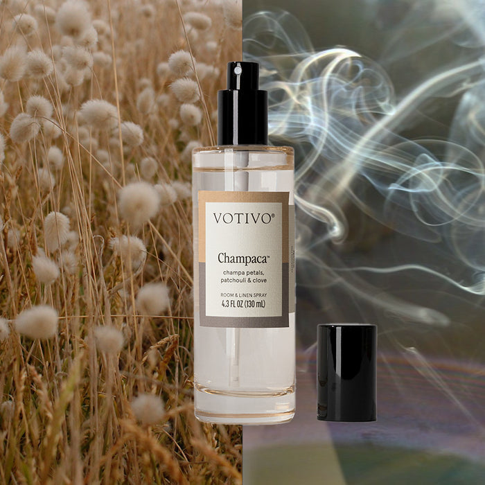 A clear Votivo Champaca Room & Linen Spray bottle with a black spray top is displayed against a split backdrop of soft wheat grass on one side and swirling smoke on the other, with its cap placed beside it.