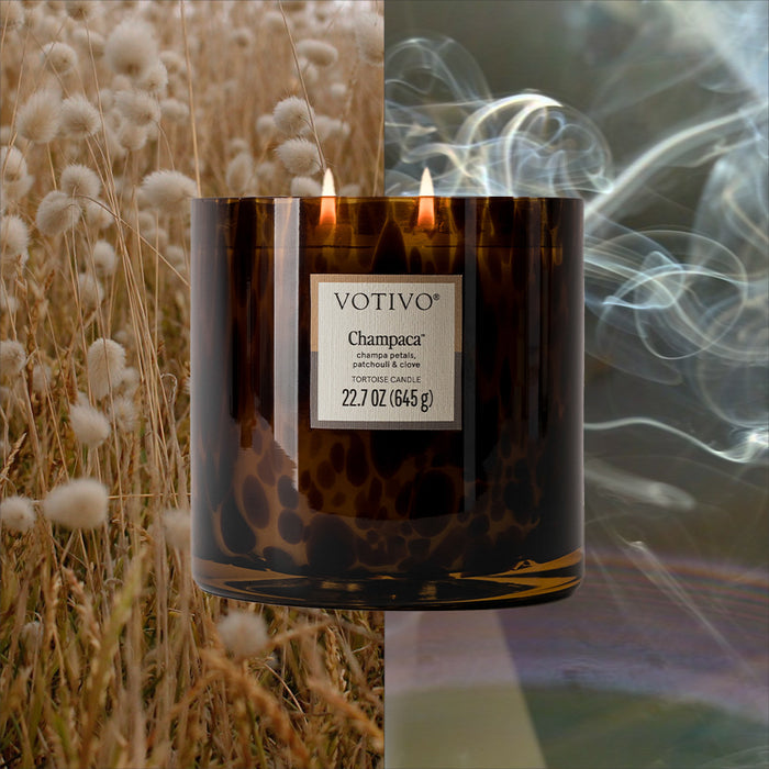 A Votivo Champaca Tortoise Candle with two flames sits at the center of a split background: one half features a sunlit field with white flowers, while the other half shows swirling smoke. Made from soy blend wax for a clean burn.