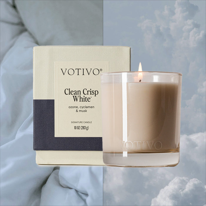 A Votivo Clean Crisp White 10 oz. Signature Candle glows in a glass jar beside its box, set against blue bedding and a cloudy sky, capturing the unique fragrance and soy-blend wax of this premium candle.
