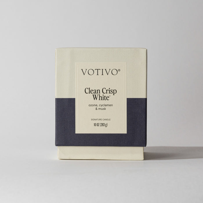The Votivo Clean Crisp White 10 oz. Signature Candle features a minimalist cream and dark gray box, labeled with scent notes of ozone, cyclamen, and musk. Made with a soy blend wax for a unique fragrance experience.