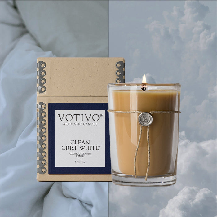 A Votivo Clean Crisp White 6.8 oz. Aromatic Candle sits lit in a glass jar beside its box on soft blue bedding, both set against a cloudy sky background.