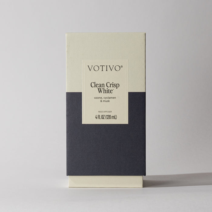 A rectangular Votivo Clean Crisp White Reed Diffuser box, featuring ozone, cyclamen, and musk scents. Its minimalist cream and dark gray design with fiber reeds ensures a continuous fresh fragrance against a light background.