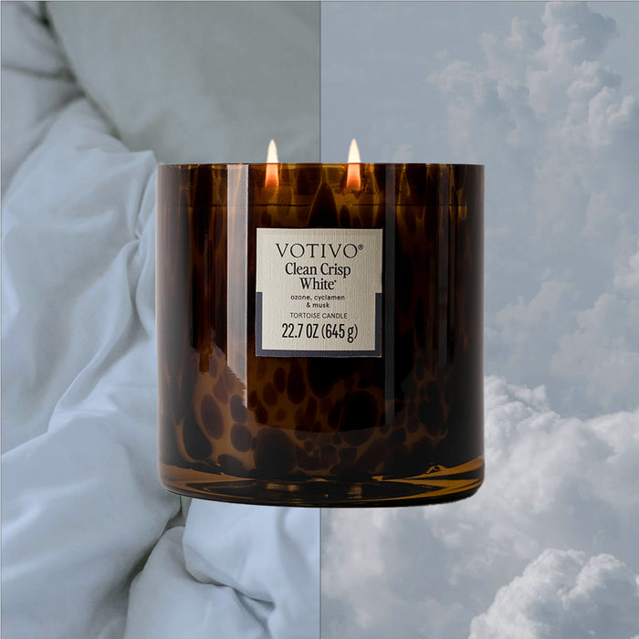 The Votivo Clean Crisp White Tortoise Candle, in brown glass with two flames, sits before a backdrop of white bedding and a cloudy sky, evoking its fresh fragrance and peaceful ambiance.