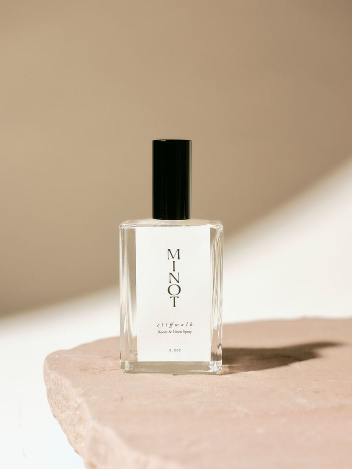 Clear glass bottle of MINOT Cliffwalk room and linen spray with black cap, placed on a textured stone surface, highlighted by soft natural light.
