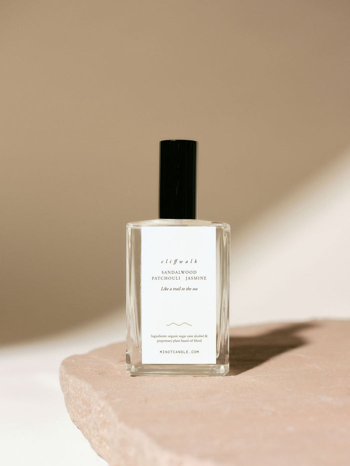 A bottle of MINOT's Cliffwalk room and linen spray with sandalwood, patchouli, and jasmine scents sits on a stone surface, with a minimalistic design and soft lighting casting gentle shadows.