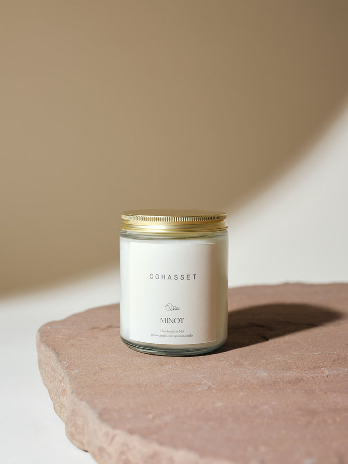 A non-toxic soy candle in a glass jar with a gold lid labeled "COHASSET" placed on a smooth, round stone surface against a neutral background.