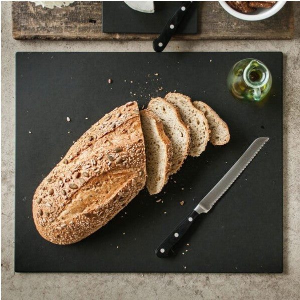 Top-down view of Cut & Serve Board showing black rectangular board with sliced multigrain loaf, serrated knife, and olive oil bottle on a stone countertop