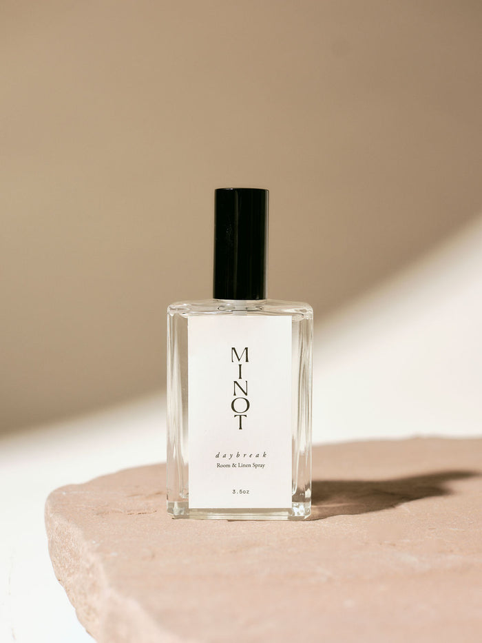Clear glass bottle of MINOT Daybreak room and linen spray with black cap, placed on a textured stone surface, highlighted by soft natural light.