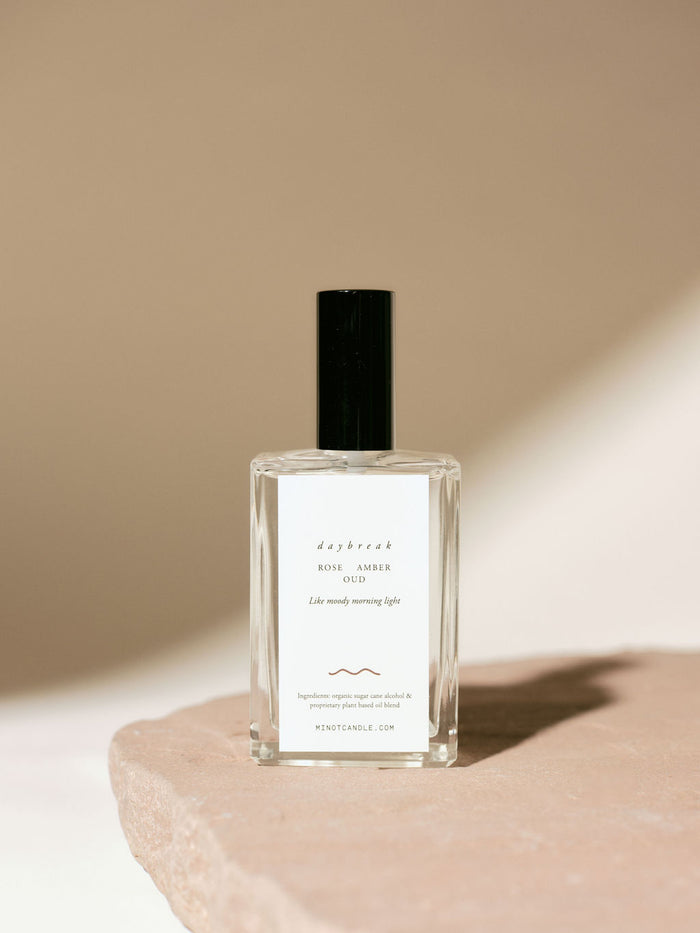 A bottle of MINOT's Daybreak room and linen spray with rose, amber, and oud scents sits on a stone surface, with a minimalistic design and soft lighting casting gentle shadows.