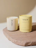 A non-toxic soy candle in a glass vessel labeled "MINOT" sits on a textured stone surface next to a yellow cylindrical box labeled "Dewpoint: Chamomile, Spruce, Amber"