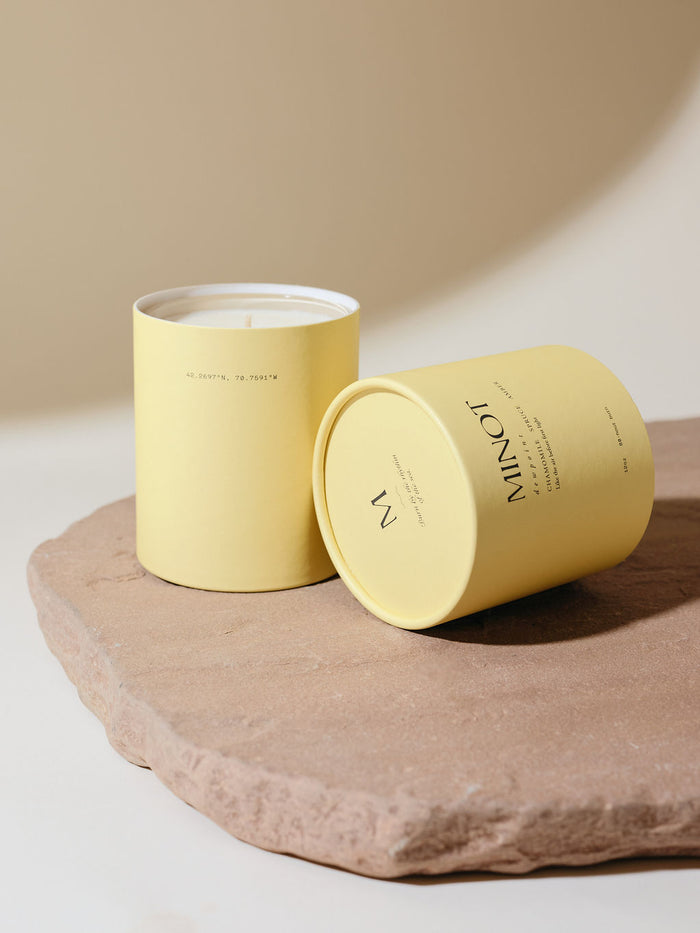 Yellow cylindrical packaging holds a MINOT Dewpoint scented candle with its lid tilted to the side on a textured stone slab. The background is a soft beige color.