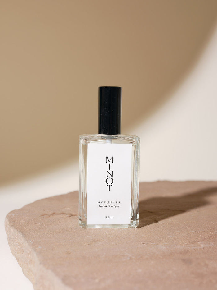 A 3.5oz glass bottle of MINOT Dewpoint Room & Linen Spray sits on a textured stone surface. The label is white with black text, and the cap is black. The background is softly lit in neutral tones.