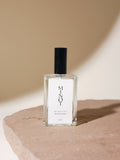 A 3.5oz glass bottle of MINOT Dewpoint Room & Linen Spray sits on a textured stone surface. The label is white with black text, and the cap is black. The background is softly lit in neutral tones.