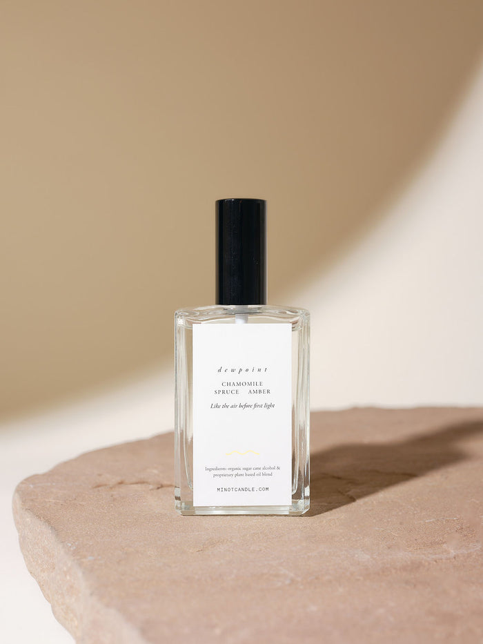 A clear glass bottle of MINOT Dewpoint Room & Linen Spray, labeled "Chamomile, Spruce, Amber" and "Like the air before first light" on a textured stone surface, with soft shadows in the background.