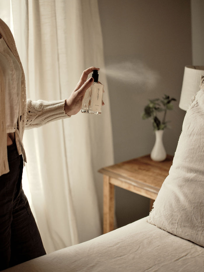 Person spraying MINOT room and linen spray in a cozy bedroom with neutral decor, wooden side table, and white vase with greenery.