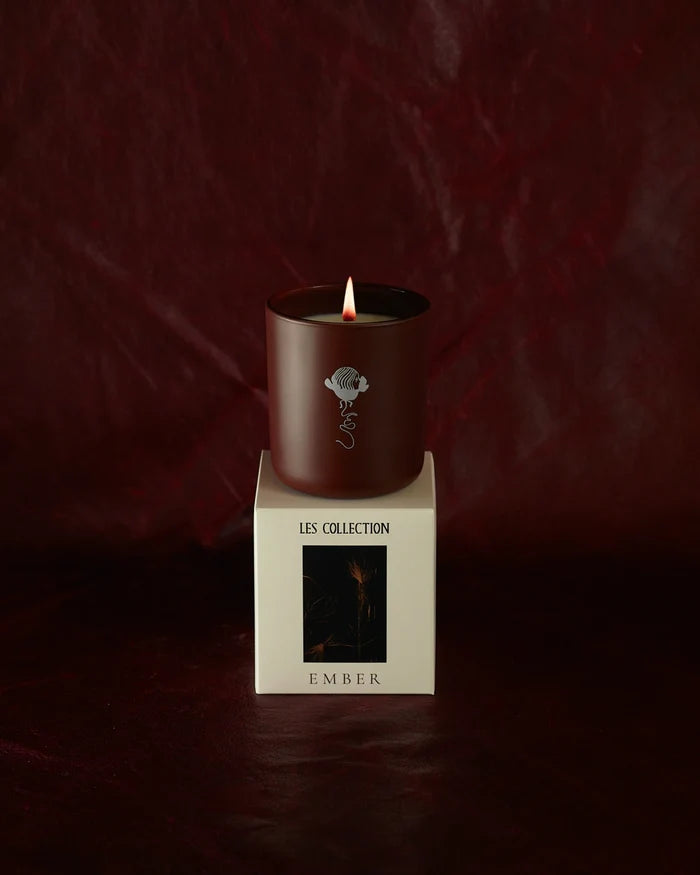 A lit LES Collection Ember candle in a dark red vessel sits on a white box labeled "LES Collection Ember." The background is a rich, dark red.