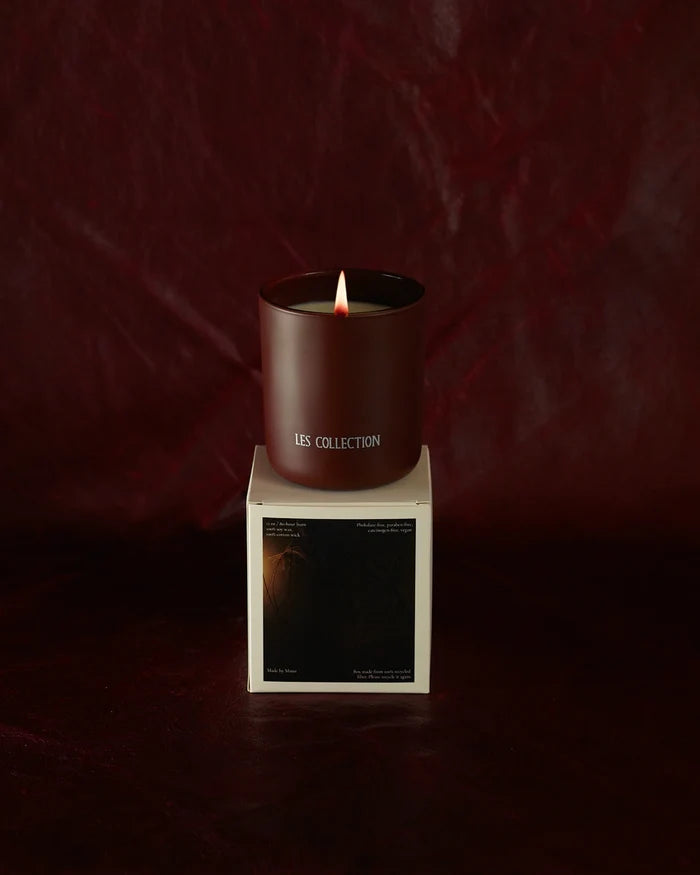 A lit LES Collection Ember candle in a dark red vessel labeled "LES Collection" sits atop a white box. The background is a rich burgundy.