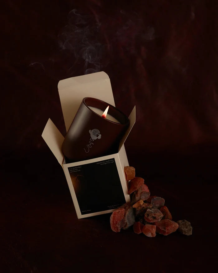 A lit LES Collection Ember candle in a dark red vessel sits partially inside an open box. Smoke rises from the flame. Rough red and gray stones are scattered beside the box against a dark background.